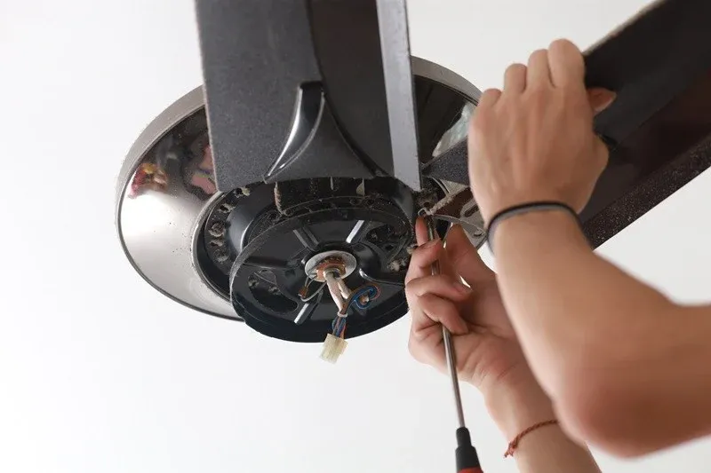 A person's hands are shown working on the underside of a ceiling fan, using a screwdriver to make adjustments.