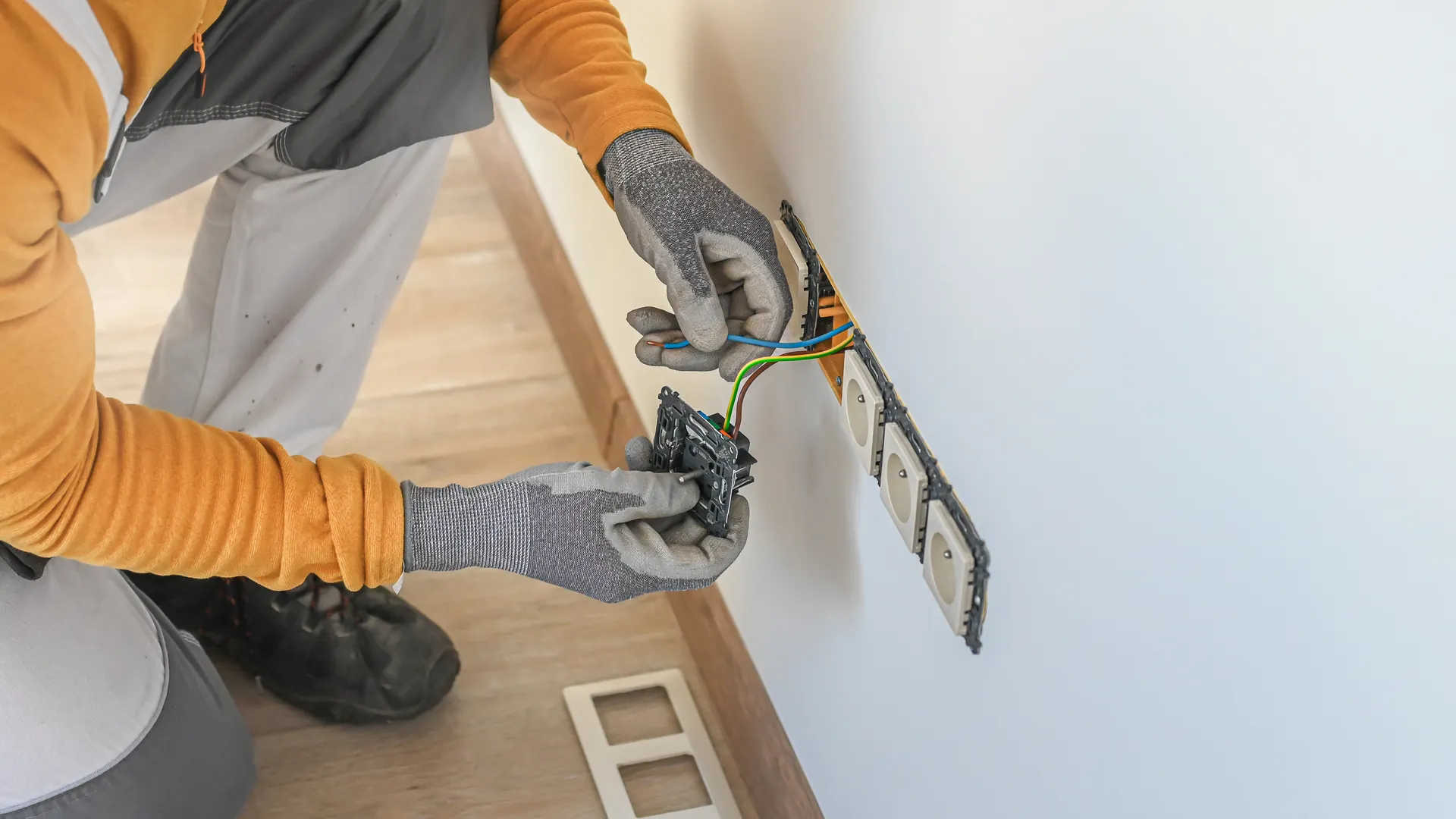 A person in gloves is working on electrical wiring near a wall outlet.