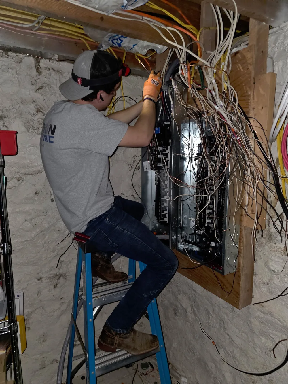 A person on a ladder working on electrical wiring in a basement.
