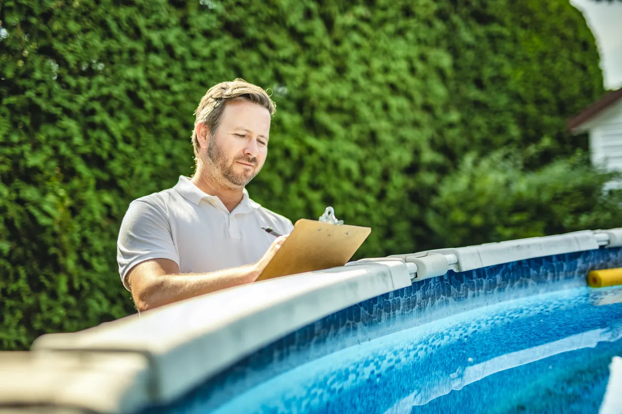 A man in a polo shirt stands by a swimming pool, holding a clipboard and smiling.