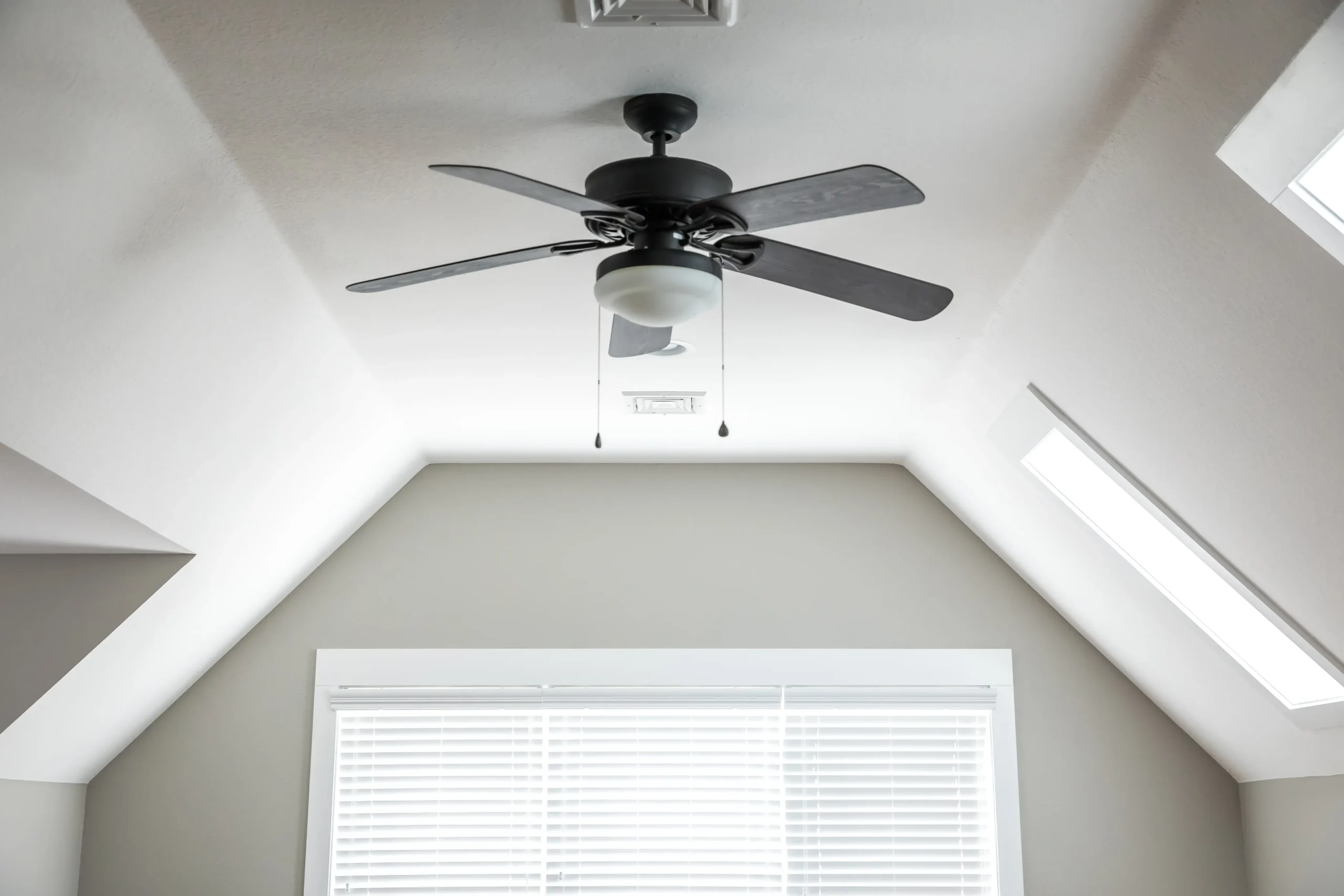 A ceiling fan with five blades is mounted on a sloped ceiling, above a window with white blinds.