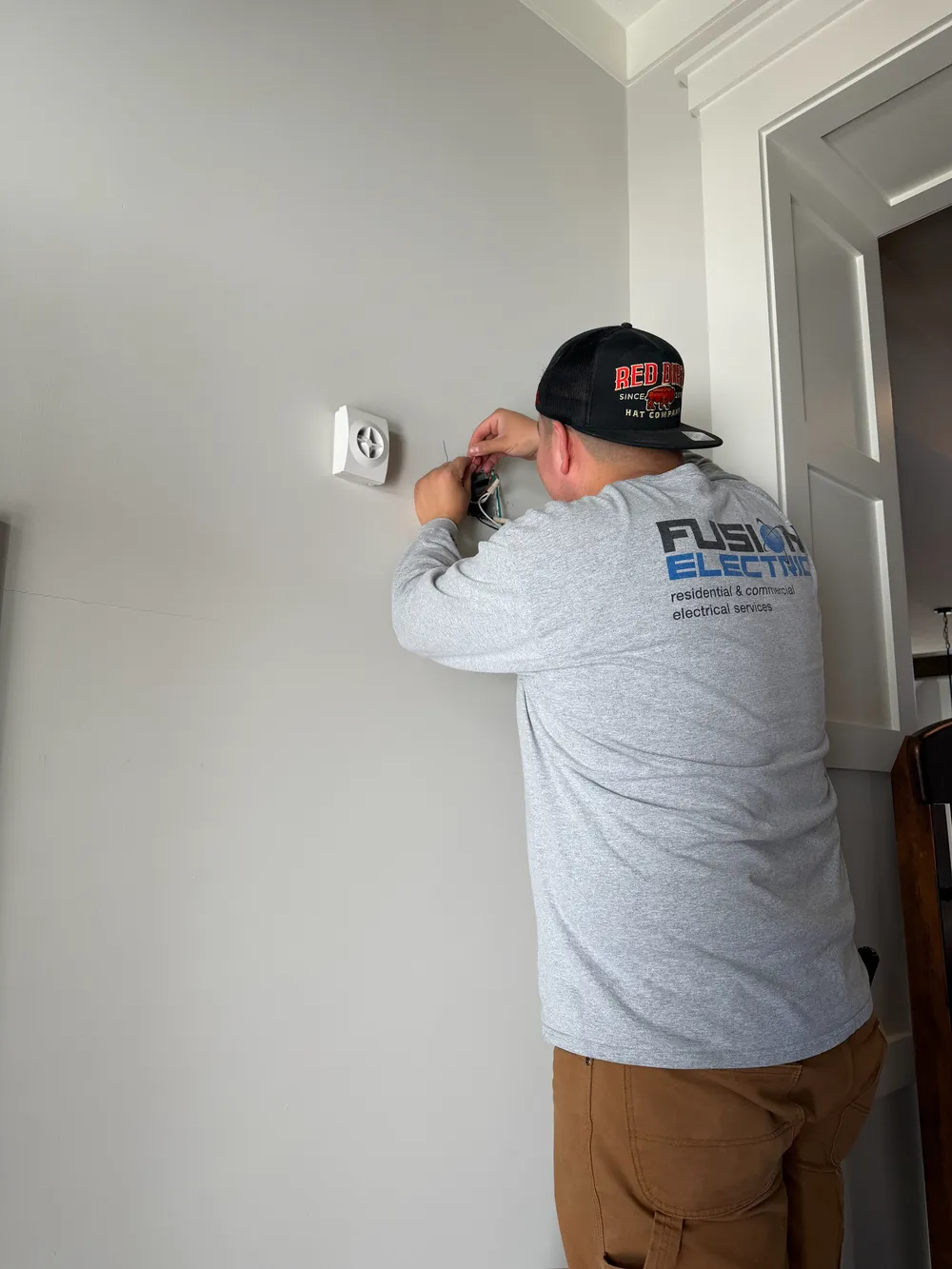 A person wearing a gray long-sleeve shirt and a cap is working on an electrical installation on a wall.