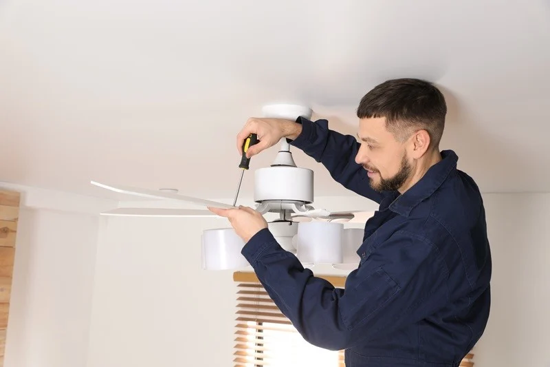 A man in a blue jumpsuit is installing or repairing a ceiling fan with a screwdriver.