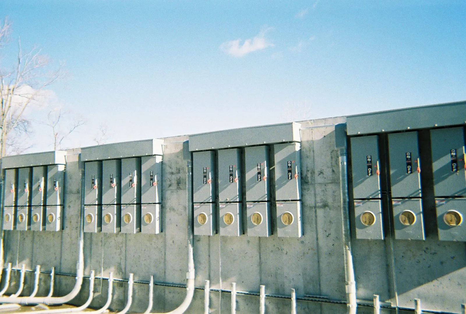 A row of utility meters mounted on a concrete wall, with pipes extending from the bottom. The sky is clear with a few clouds.