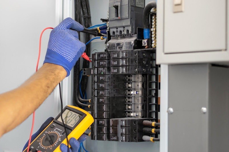 A close-up of a person's hand wearing a blue glove, using a multimeter on a circuit breaker panel.