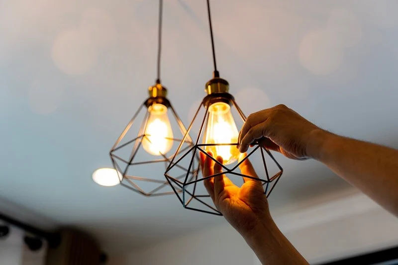 A person adjusting a light bulb in a geometric wire pendant light fixture.