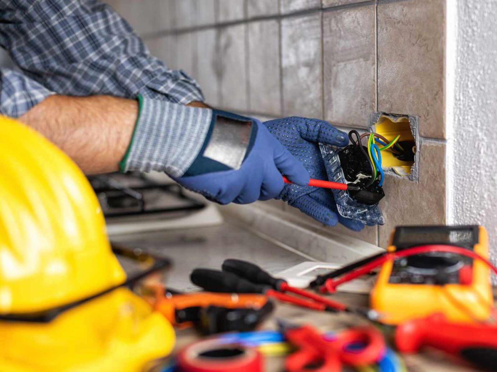 A person wearing blue gloves and a plaid shirt is working on electrical wiring at a wall outlet, with tools and a hard hat nearby.