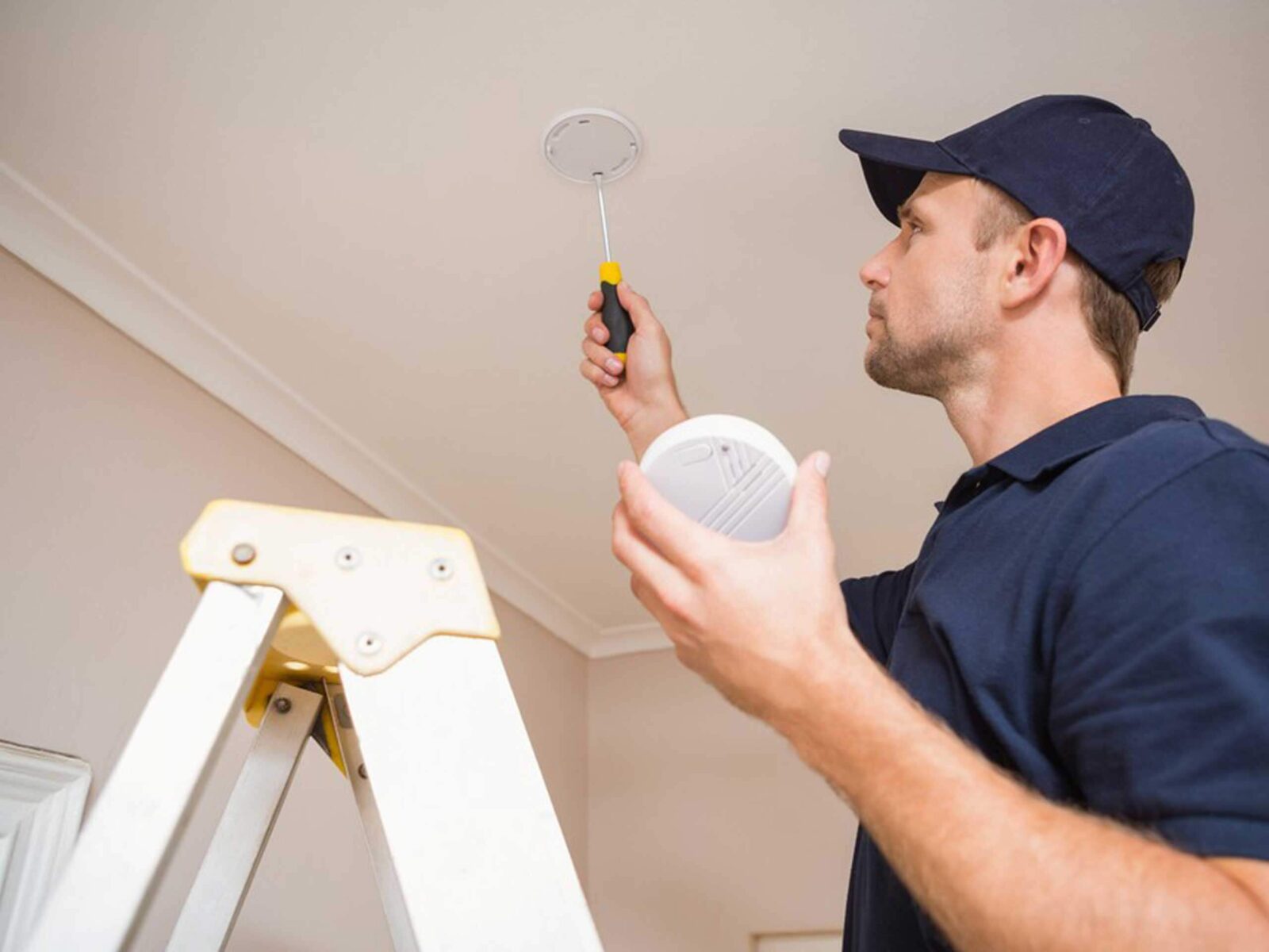 A person in a cap standing on a ladder, installing a smoke detector on the ceiling.