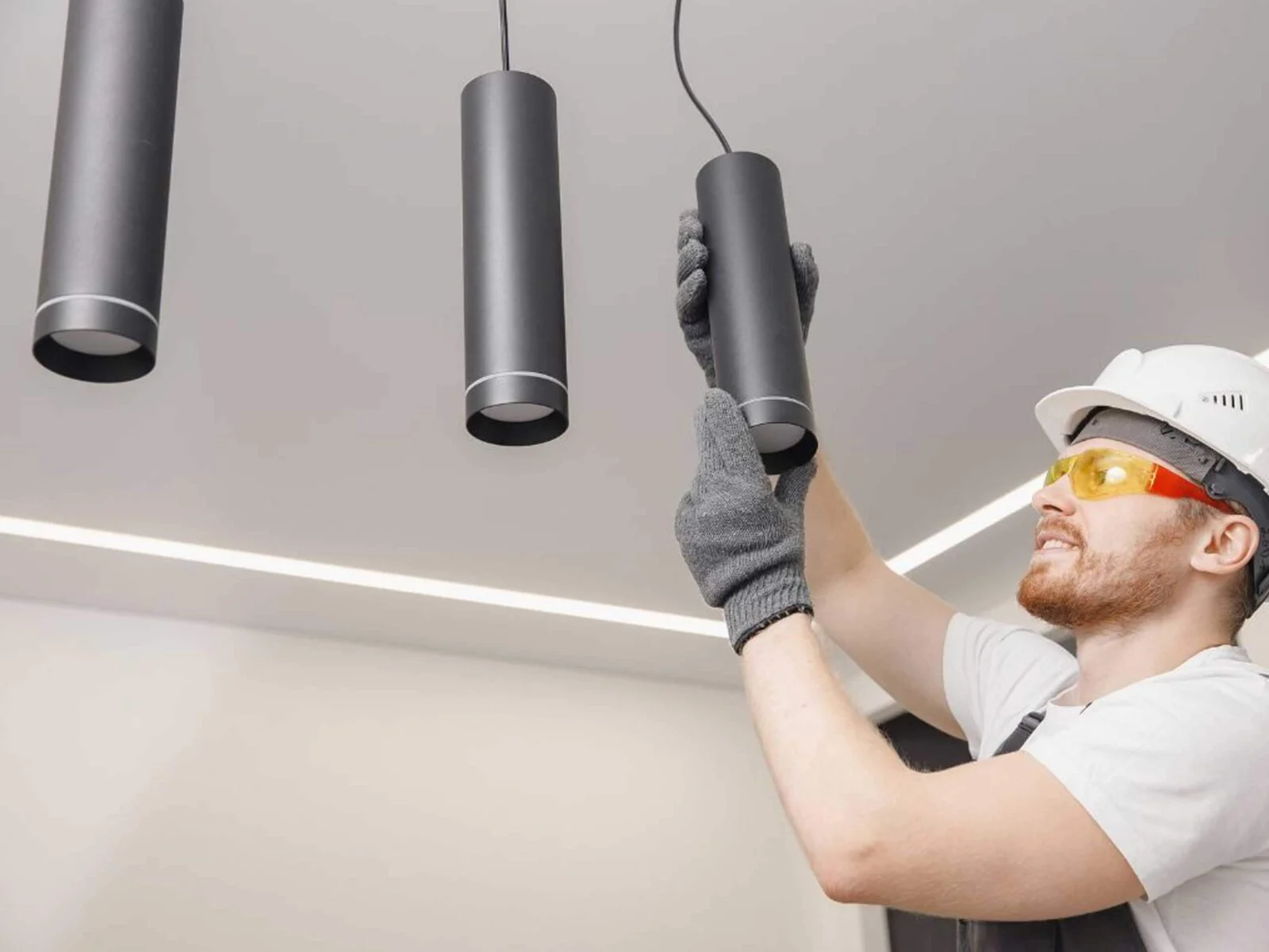 A worker wearing a hard hat and safety glasses is installing black cylindrical pendant lights in a ceiling.