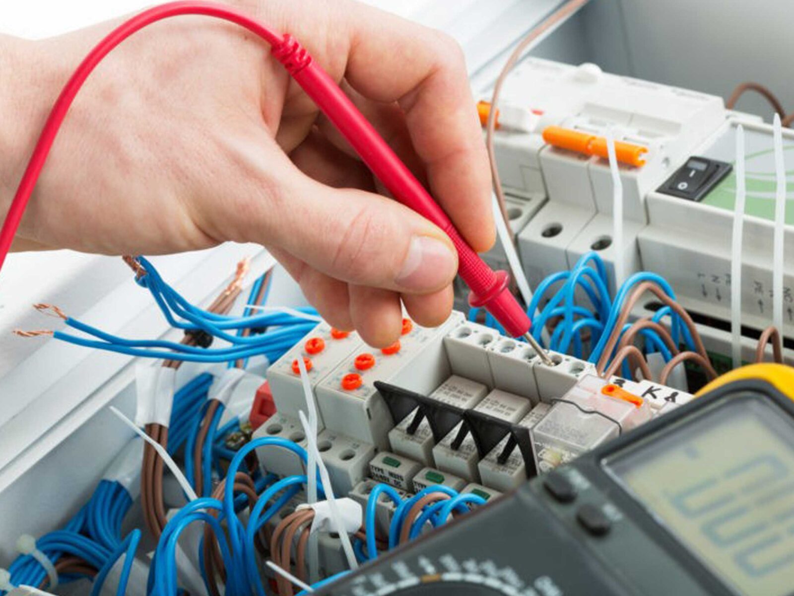 A hand holding a red testing probe is measuring electrical components among a tangled array of wires, circuit breakers, and a multimeter in an electrical panel.