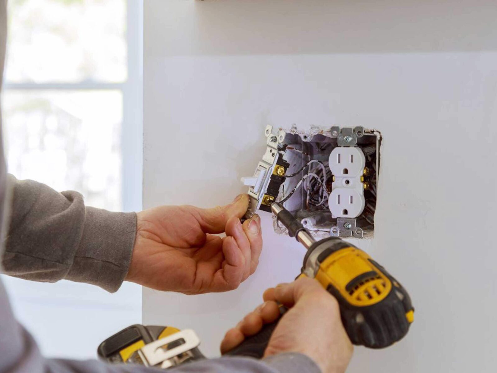 A person using a power drill to install or repair an electrical outlet on a wall.