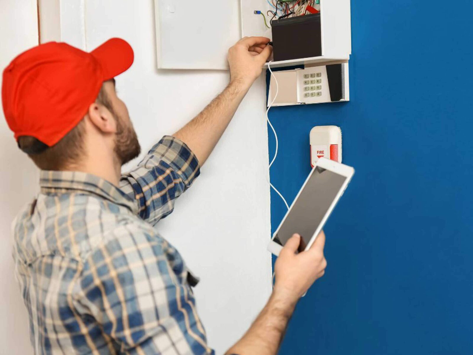 A man in a red cap and plaid shirt works on a security system, holding a tablet in one hand and adjusting wires in a box on the wall.