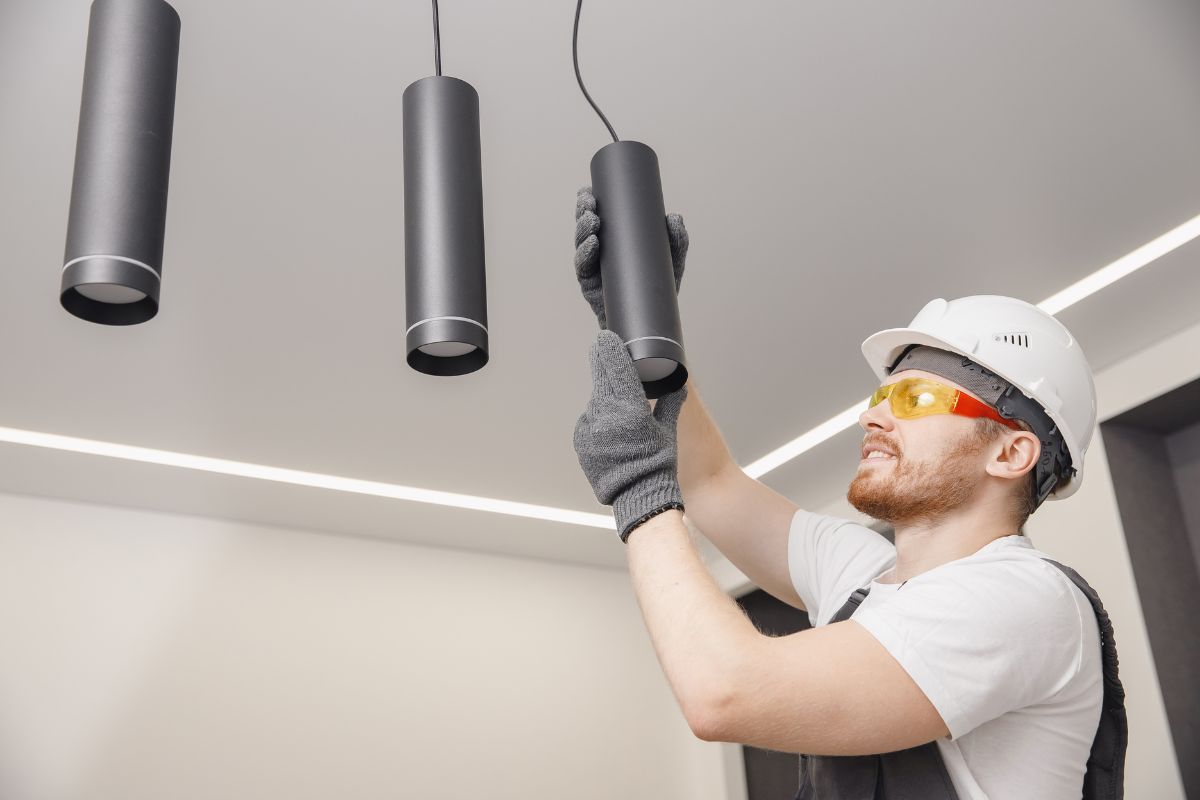 A man wearing a hard hat and safety goggles is installing hanging light fixtures in a modern interior.
