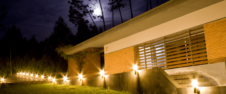 A modern house at night, illuminated by pathway lights, with a moonlit sky and trees in the background.