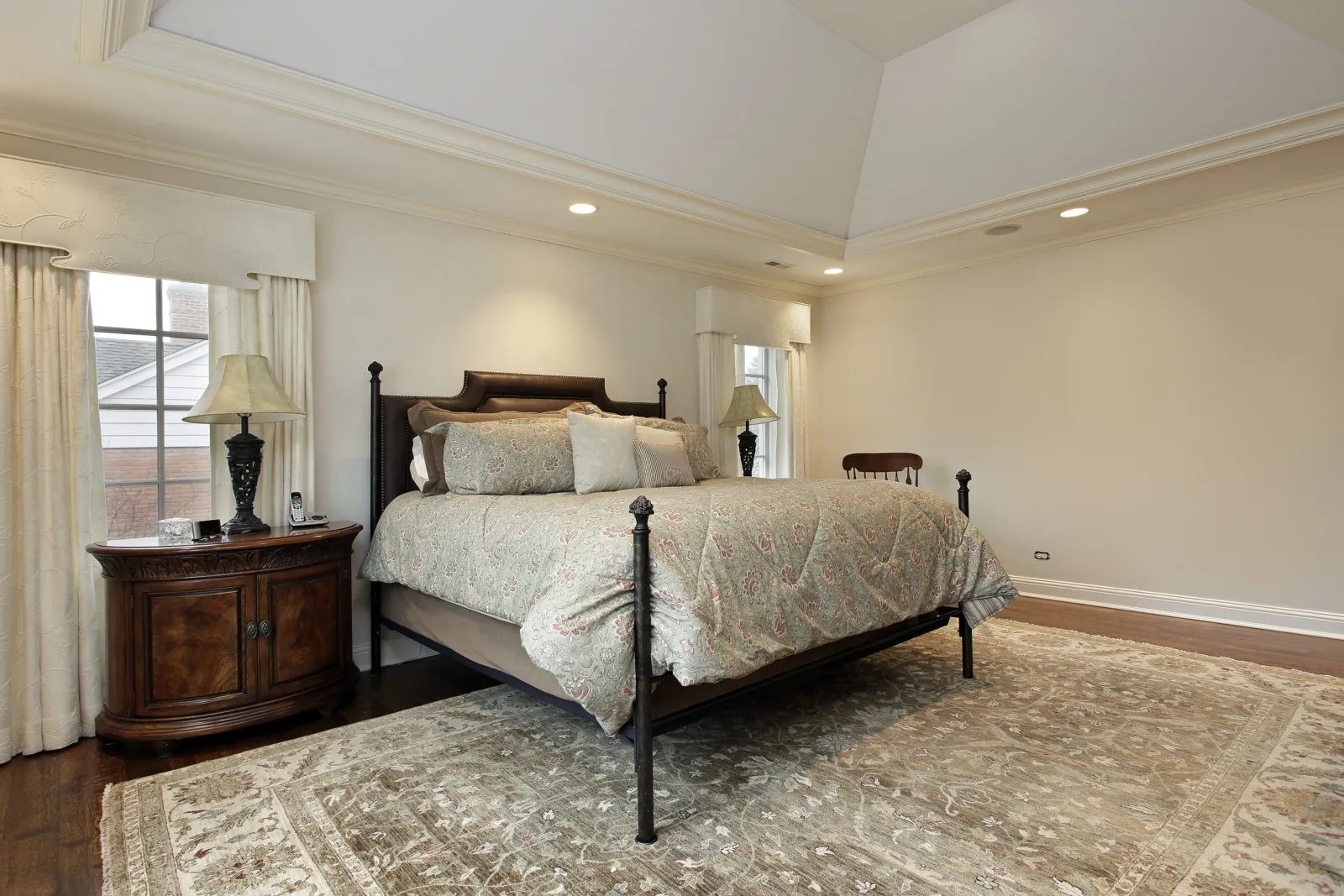 A spacious bedroom featuring a large bed with an elaborate comforter, bedside tables with lamps, and a plush area rug on hardwood flooring.