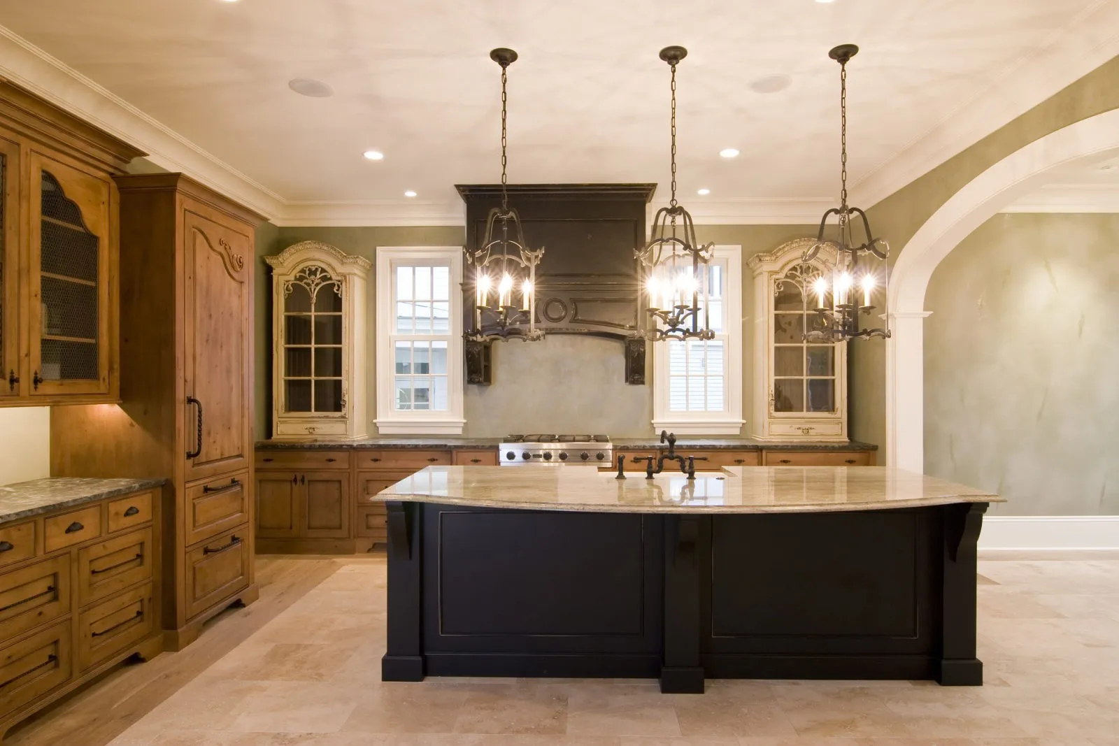A spacious kitchen featuring a large central island with a dark base and a light-colored countertop, wooden cabinetry with glass fronts, and elegant hanging light fixtures.